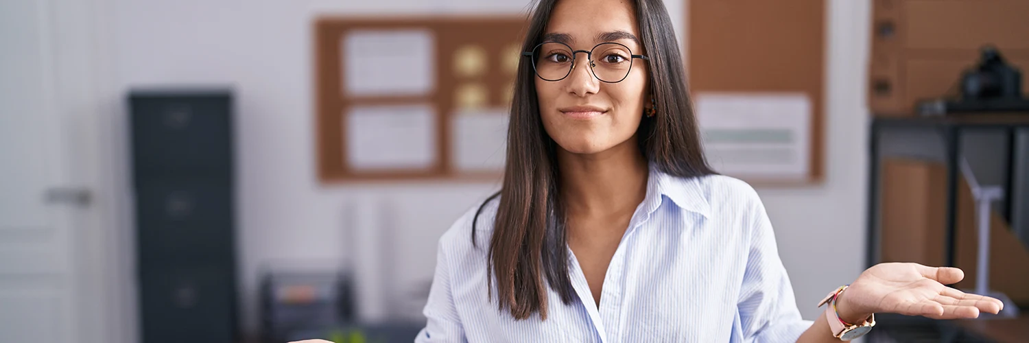Junge Frau mit Brille hebt fragend eine Hand in einem modernen Büro.