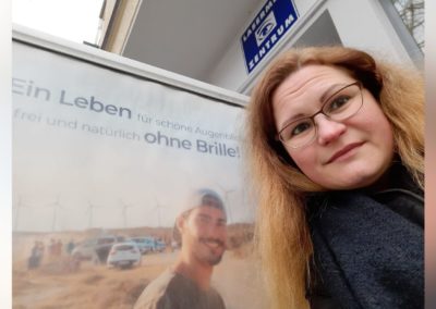 Blonde Frau mit Brille steht neben Plakat vor dem Augenlaser-Zentrum in der Schönhauser Allee